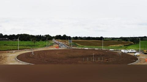 An aerial image of Newby West roundabout while work is ongoing. The roundabout is isolated from a temporary road where traffic is flowing by concrete blocks. The middle of the roundabout is covered in soil, with some thin posts on three areas of it. There are white vehicles associated with the roadworks parked next to the roundabout on what appears to be a dirt track. Fields surround the A595 road which now leads to a temporary road.