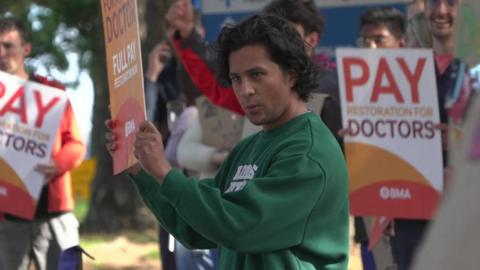 Doctors stand outside a hospital carrying placards calling for pay restoration during a strike organised by the British Medical Association. The main person in shot is wearing a green Adidas jumper.