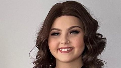 Molly Cuddihy, a young woman with curly, long, dark hair and a nose ring, smiles broadly at the camera. 