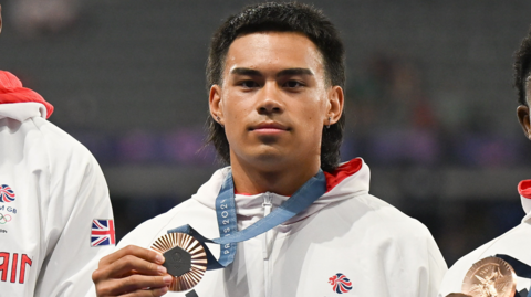 Louis Hinchliffe, wearing Team GB's white 2024 Olympics jacket, holds up his bronze medal in Paris
