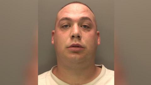 A police photo of a man with a buzz cut and clean shaven looking into the camera. He is wearing a white t-shirt.