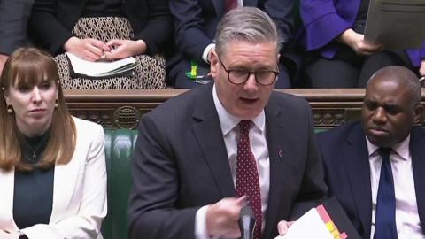 A man in a dark suit, white shirt and red tie with glasses stands at a lecturn speaking across to someone. A red haired woman and a man in a navy suit sit on green benches next to him. 