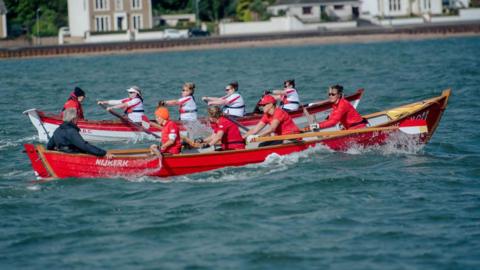 Two coastal rowing teams go head to head one in a red boat and red tops, the other in a white boat with white tops and a red stripe