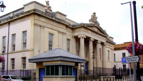 Shows a large court building with four pillars at the front and a security hut at the left with a silver-coloured car beside it.