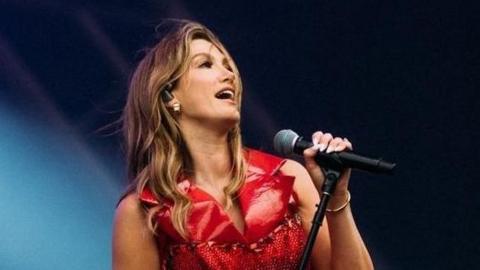 Delta performing on stage in Ormeau Park in Belfast, she is wearing a red dress and red thigh high boots, she is gripping the microphone and leaning forward singing