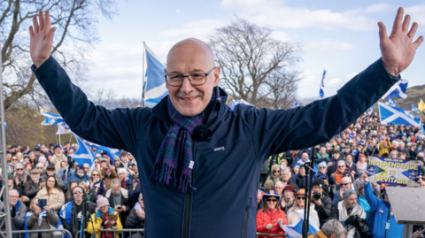 Swinney standing on a stage with arms raised in front of a large outdoor crowd holding Scottish flags, with banners and signs visible among the audience and trees in the background.
