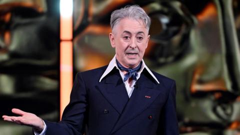 Alan Cumming on stage in a black suit talking at the Bafta Film Awards in London, 2026