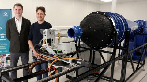 A man in a black blazer and suit shirt standing next to a man in jeans and a navy shirt. They are standing next to what looks like an F1 exhaust.