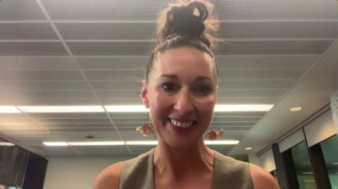 Rachel Duffy. She has long brown hair tied up in a bun. She is wearing dangly earrings and a khaki sleeveless top.