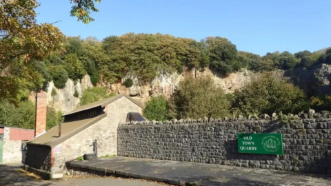 A stone wall with a green sign that reads 'Old Town Quarry' next to a stone house and lots of trees in the background