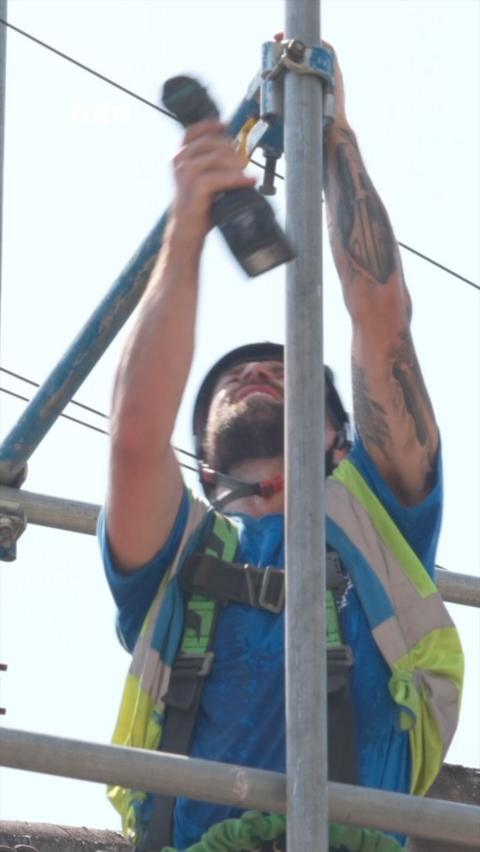 A man wearing a yellow high vis jacket and a safety helmet stares upwards as he works on some metal scaffolding