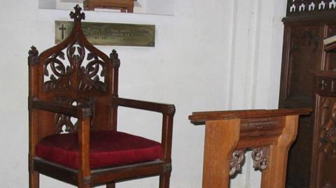 Ceremonial seat made from oak with seat cushion covered in red velvet. 
The chair is cut ornately and has a cross on the top. It is placed against a white wall of the church  with a small lectern in front.