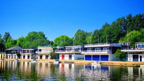 A row of brightly coloured houses along a waterside, with green trees behind and bright blue sky above