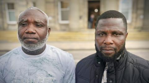 Two men shown side-by-side in a split portrait, both facing the camera outdoors in front of a blurred building. The man on the left has a short haircut with a grey beard and is wearing a light-coloured patterned shirt, while the man on the right has short hair, a trimmed beard, and is wearing a dark quilted jacket over a shirt.