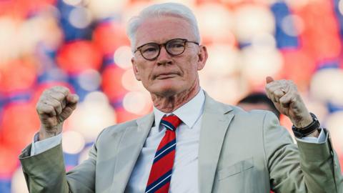 Eamonn McManus, wearing a beige suit jacket, light coloured shirt and red and blue tie, pumps both his fists in the air to celebrate after a St Helens game