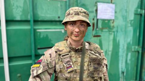 Laura Holloman standing in front of a green shipping container, wearing army camouflage and bucket hat. She has green paint on her face