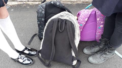 There are a pair of girls' legs in white socks and black patent shoes, and another pair of girls' legs in black leggings and black lace up boots. The feet are stood by three children's backpacks