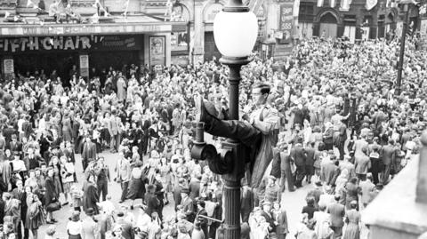 Crowds celebrate VE Day and mad sit on lamp post.