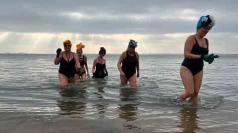 Five women walk out of the sea, four are wearing swimsuits and one is in a bikini. Three of them are wearing flower head-dresses, and another three are wearing gloves. You can see the sun's rays poking through the clouds behind them