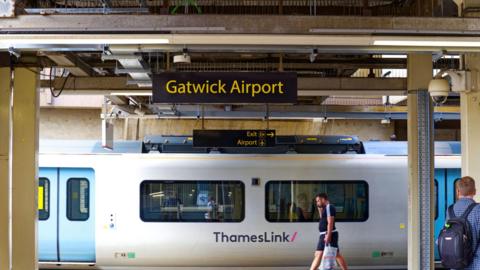 Sign at Railway station with platform and leaving white train of Thameslink at Gatwick Airport London on a cloudy summer day.