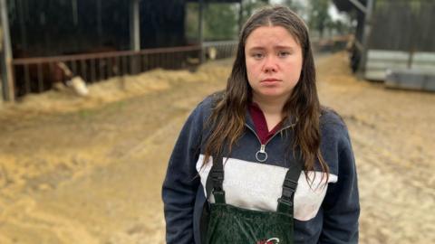 A young farmer stands in a yard with cattle sheds in the background. There are cows and young calves eating hay in the sheds. 