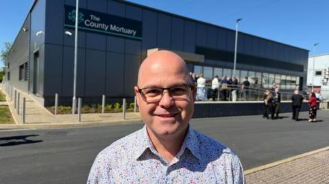 A man in a multi-coloured spotted shirt stands in front of a large black building. "The County Mortuary" is written on the side.