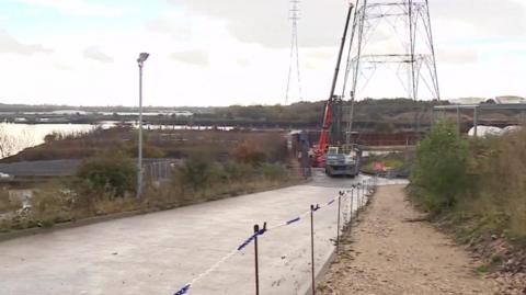 View of the entrance to the Howdon treatment works, with the banks of the River Tyne to the left, a road with a digger at the end of it, next the the lower part of an electricity pylon.