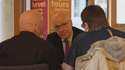 John Swinney faces the camera while in conversation with a man and a woman, Ian and Fiona Inglis who are facing away from the camera. Swinney is wearing a suit, shirt and tie. He is bald and is wearing glasses. From the back, Ian Inglis is bald and wearing a black shirt. Fiona has short brown hair and is wearing a navy top.