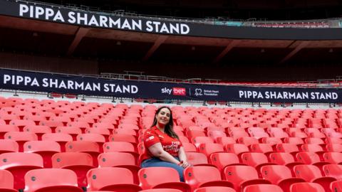 A picture of Pippa Sharman, wearing a red Every Minute Matters T-shirt, sat in Wembley stadium with "Pippa Sharman Stand" behind her on screens running behind her.
