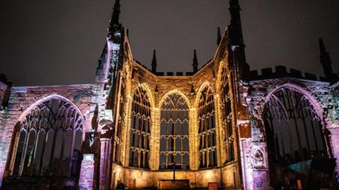 A cathedral ruins lit in golden and purple light. A cross is in the centre of the picture in a ruined chancel.