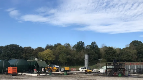 The drilling site at Horsehill in Surrey, viewed from a distance, with two cylindrical green tankers, equipment and vehicles, and a line of trees in the background.