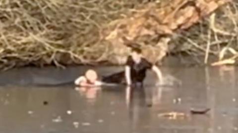 Two men at a frozen body of water - one is in the water with his arms on the ice. The other is sitting on the the ice with his legs out sideways and his torso upright with his hands on the ice. There are near a tree-lined bank.