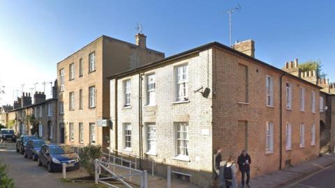 View of the Willow Walk building, in a Georgian style with big sash windows. Three people with their faces blurred are talking outside. There's some cars parked on the cobbled street.