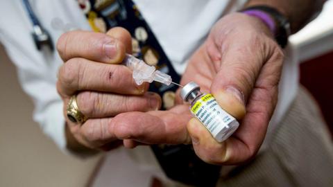 A close-up of some hands inserting a hypodermic needle into a bottle of the HPV vaccine liquid ready to be administered as an injection. 