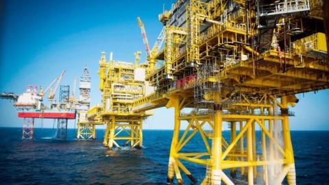 Image of an oil rig in the north sea - yellow stanchions reach down in the water from several platforms, against a blue sky and deeper blue sea
