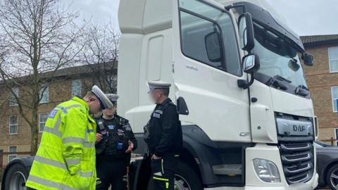 Three uniformed police officers stand next to the cab of a white lorry. The lorry is parked outside a block of flats.