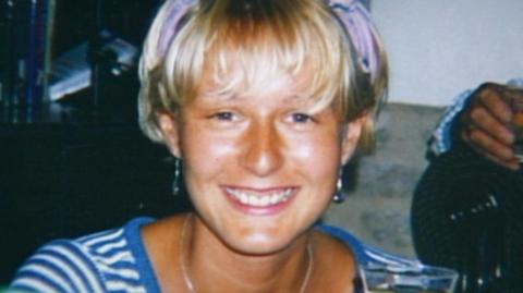 Melanie Hall is smiling at the camera wearing earrings and a blue t-shirt.