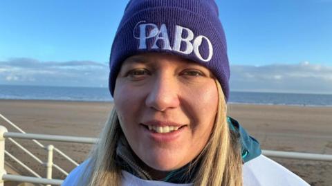 Llun pen ac ysgwyddau o Sera Mai William gyda thraeth, y môr, awyr las a chymylau gwyn y tu ôl iddi