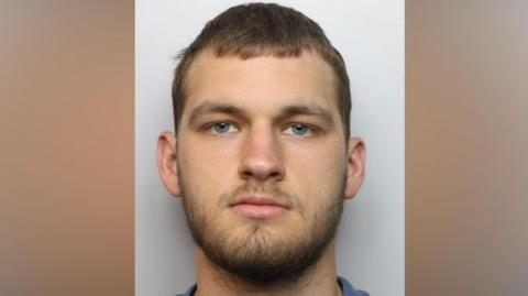 The custody photo of a man who has brown hair and a beard. He is looking directly at the camera and is wearing a blue shirt.
