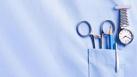 A stock image of a nurse's top pocket with a watch, pair of scissors, pencil and pen in the pocket.