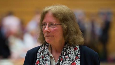 Julie Morgan looking to her right at an election count wearing a scarf with white and red flowers on it. She has light brown hair and is wearing thin-rimmed glasses