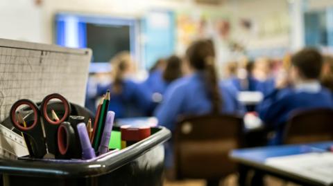 Blurred image of the backs of schoolchildren in a classroom with a box of scissors and pencils in the foreground