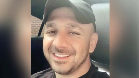 Hemawand Ali Hussein is smiling at the camera while sitting in a car. He has brown eyes and a beard and is wearing a black cap and t-shirt. 