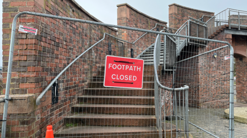 An image of a fenced off entrance to Irish Gate Bridge in Carlisle, with a red sign reading 'Footpath Closed' attached to the metal fencing. Steps, some brick and some metal, arc to the right as they climb towards the top of the bridge.