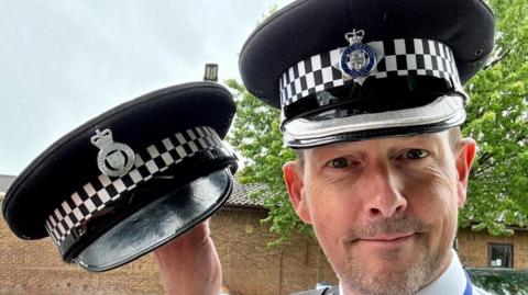 A policeman holding his lost hat up to the left of him. He is smiling and wearing his new hat also. There is a blue sky and a tree with green leaves in the background. 
