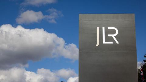 A large grey rectangular sign bearing the white JLR logo is photographed against a blue sky with scattered white clouds. A Union flag is partially visible behind the sign to the right. The image is taken from a low angle, making the sign appear prominent against the sky.