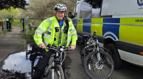 A police officer in a hi-vis jacket is on a bike and wearing a helmet. Trees and grass are in the background on the left and a police van is on the right.