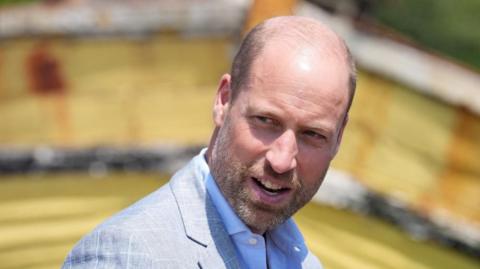Prince William in a grey jacket and blue shirt. 