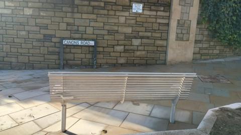 A metal bench facing a tan-coloured stone wall. The sign for Canons Road is in front of the wall, and a small 'no loading' sign can also be seen.
