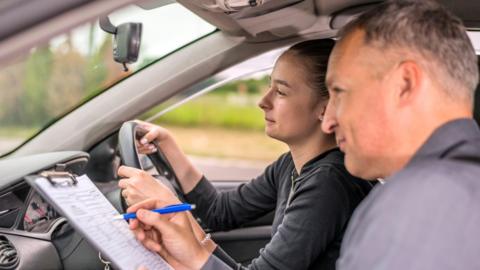 A learner driver takes a driving test with an instructor sitting in the front passenger seat, holding a clipboard and pen inside a car.
